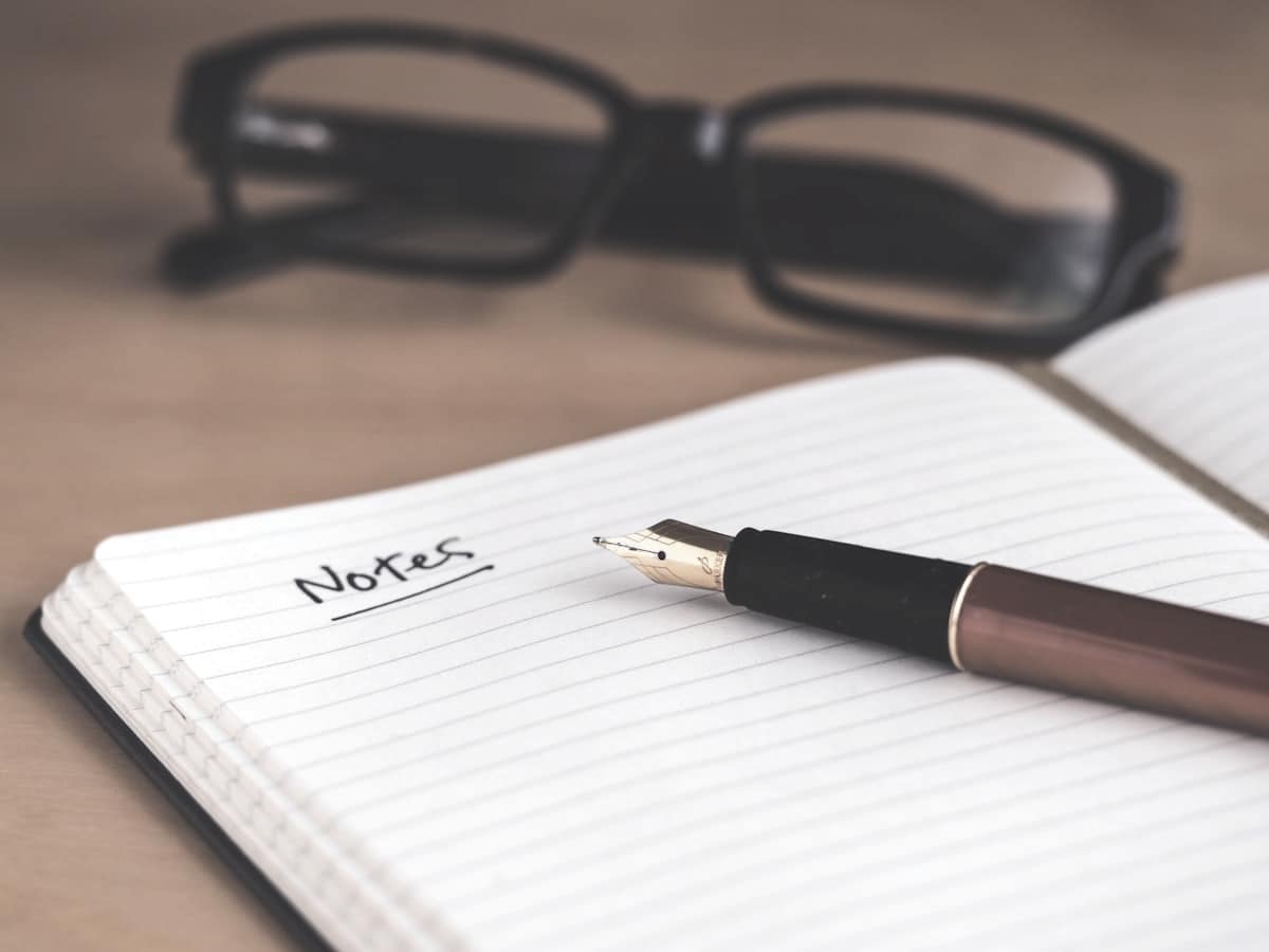 Cuaderno abierto con la palabra Notes escrita a mano, una pluma y unas gafas
